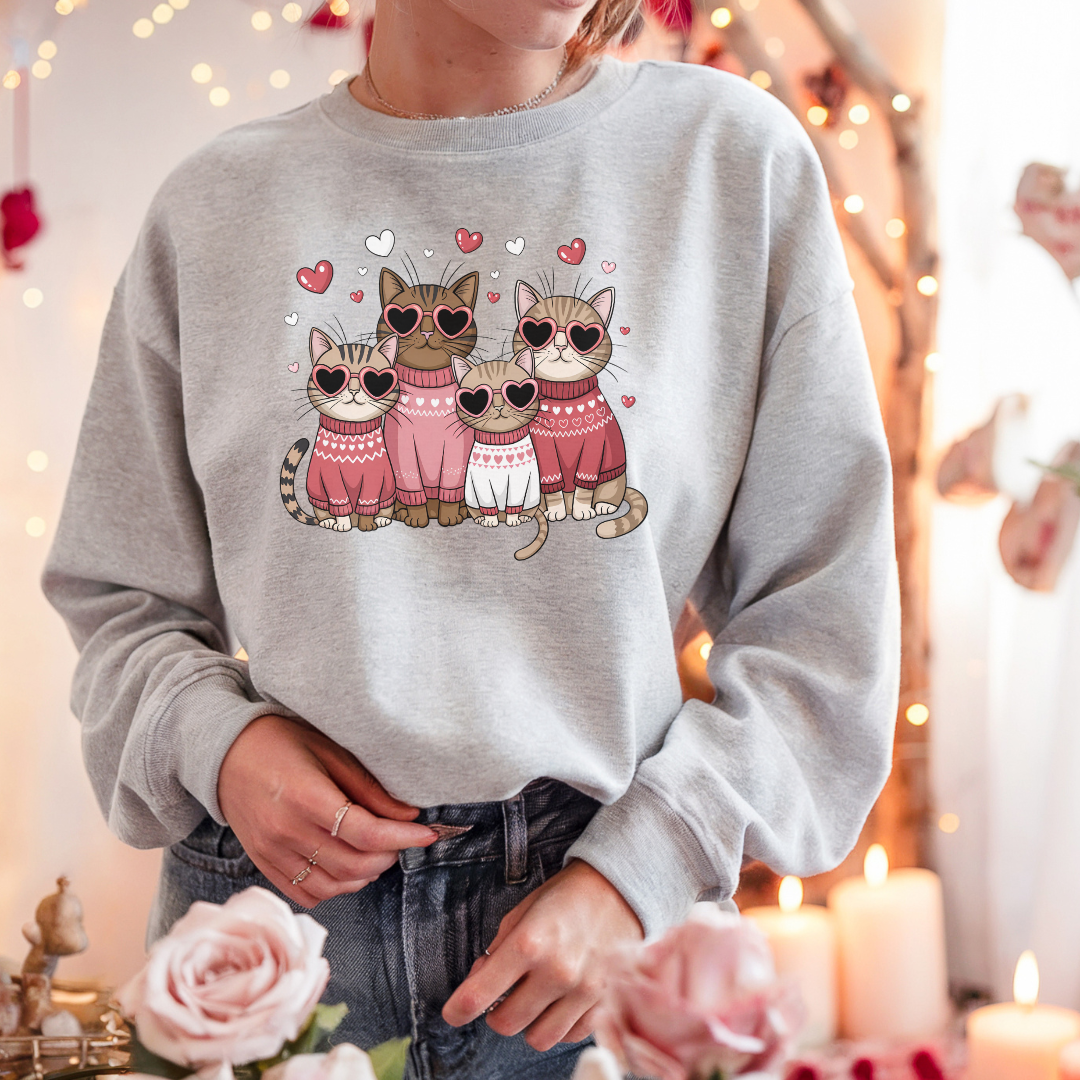  Sweatshirt gris pâle avec un motif de quatre chats portant des lunettes en forme de cœur et des pulls roses. Les chats sont entourés de petits cœurs roses.