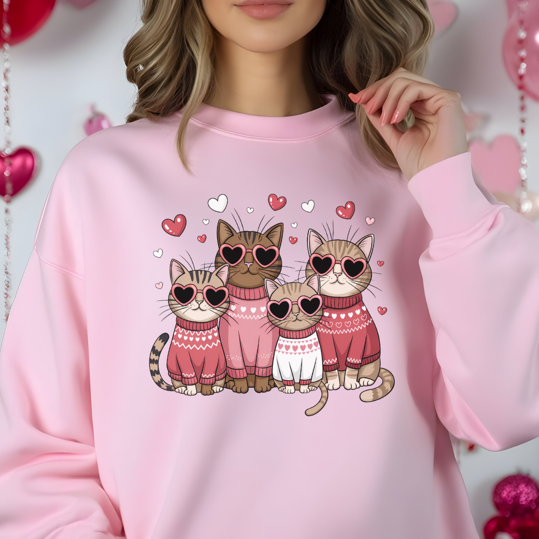  Sweatshirt rose pâle avec un motif de quatre chats portant des lunettes en forme de cœur et des pulls roses. Les chats sont entourés de petits cœurs roses.
