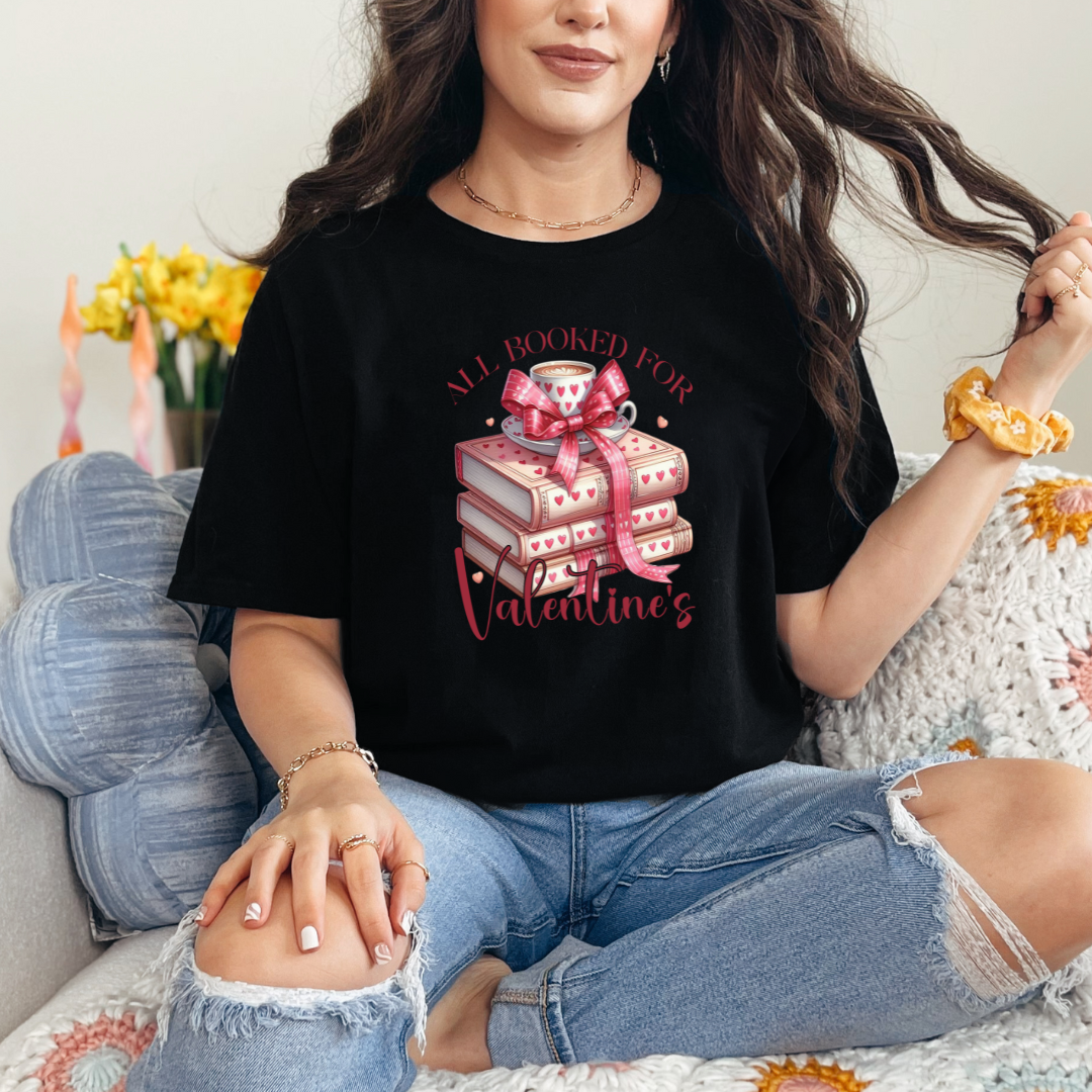 Mannequin portant un T-shirt Gildan Noir avec l’inscription “All Booked for Valentine’s”, illustré par une pile de livres décorés de cœurs et une tasse de café, style romantique et cosy.