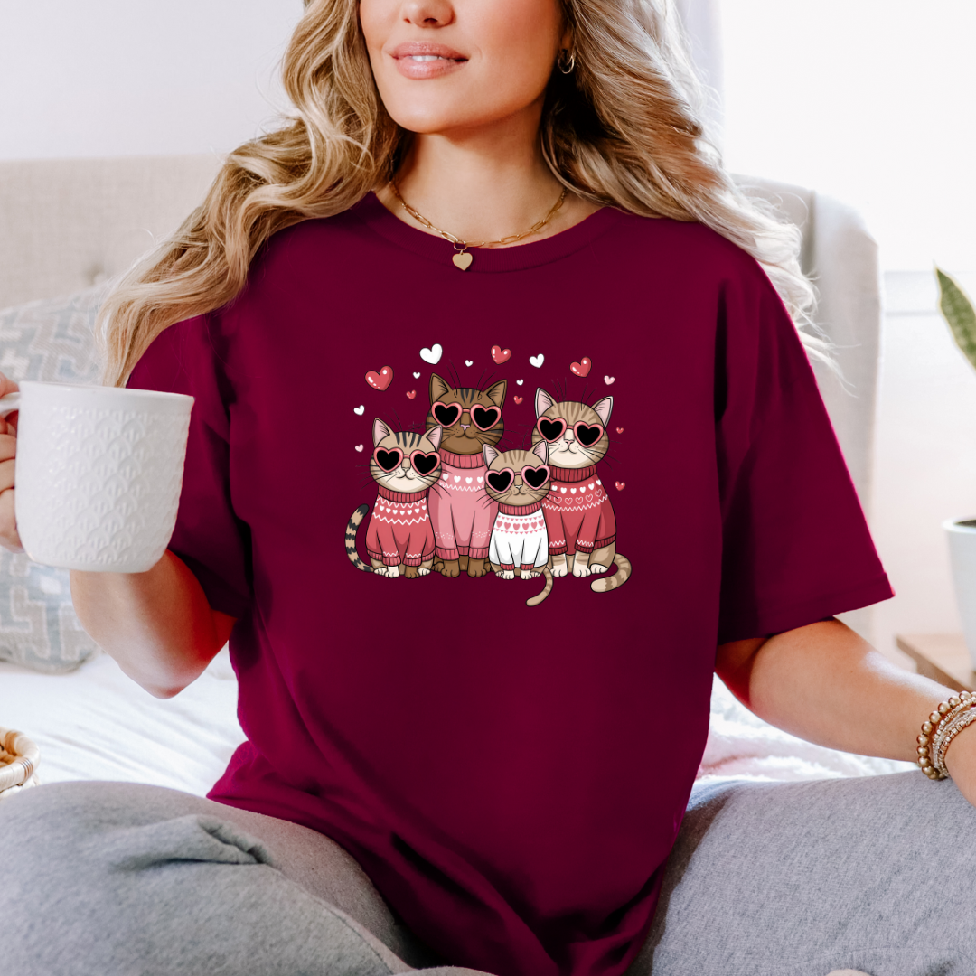 T-shirt bourgogne avec un motif de quatre chats portant des lunettes en forme de cœur et des pulls roses. Les chats sont entourés de petits cœurs roses.