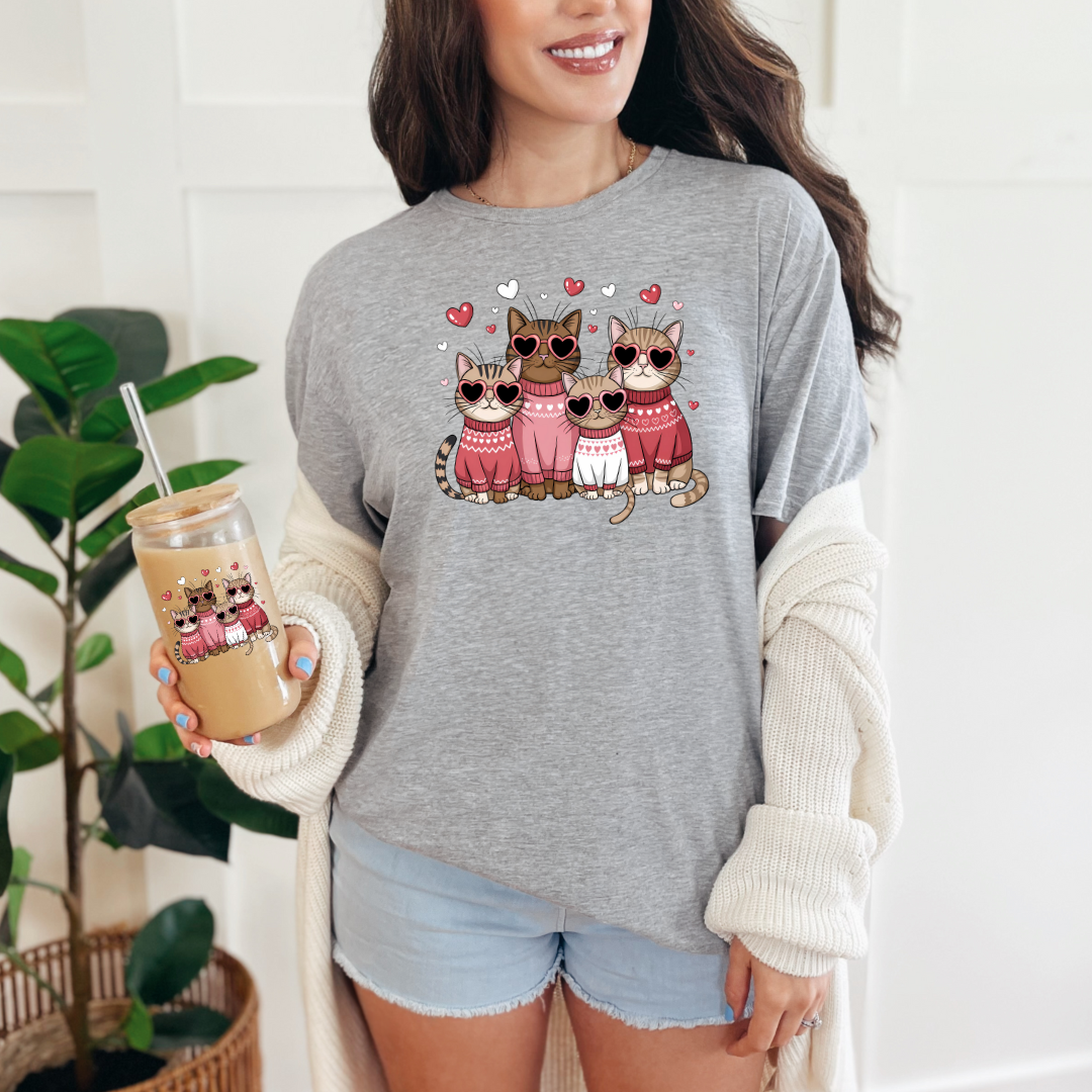 T-shirt gris pâle avec un motif de quatre chats portant des lunettes en forme de cœur et des pulls roses. Les chats sont entourés de petits cœurs roses.