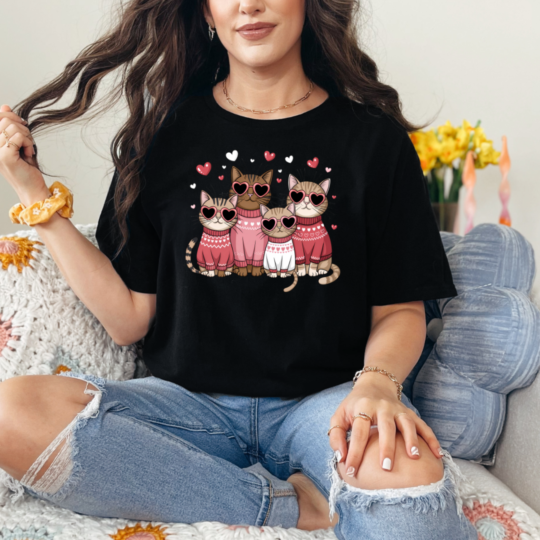 T-shirt noir avec un motif de quatre chats portant des lunettes en forme de cœur et des pulls roses. Les chats sont entourés de petits cœurs roses.