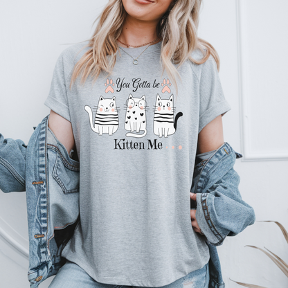 Femme portant un t-shirt noir avec une illustration de trois chats blancs dans des tasses et le texte humoristique "You Got To Be Kitten Me