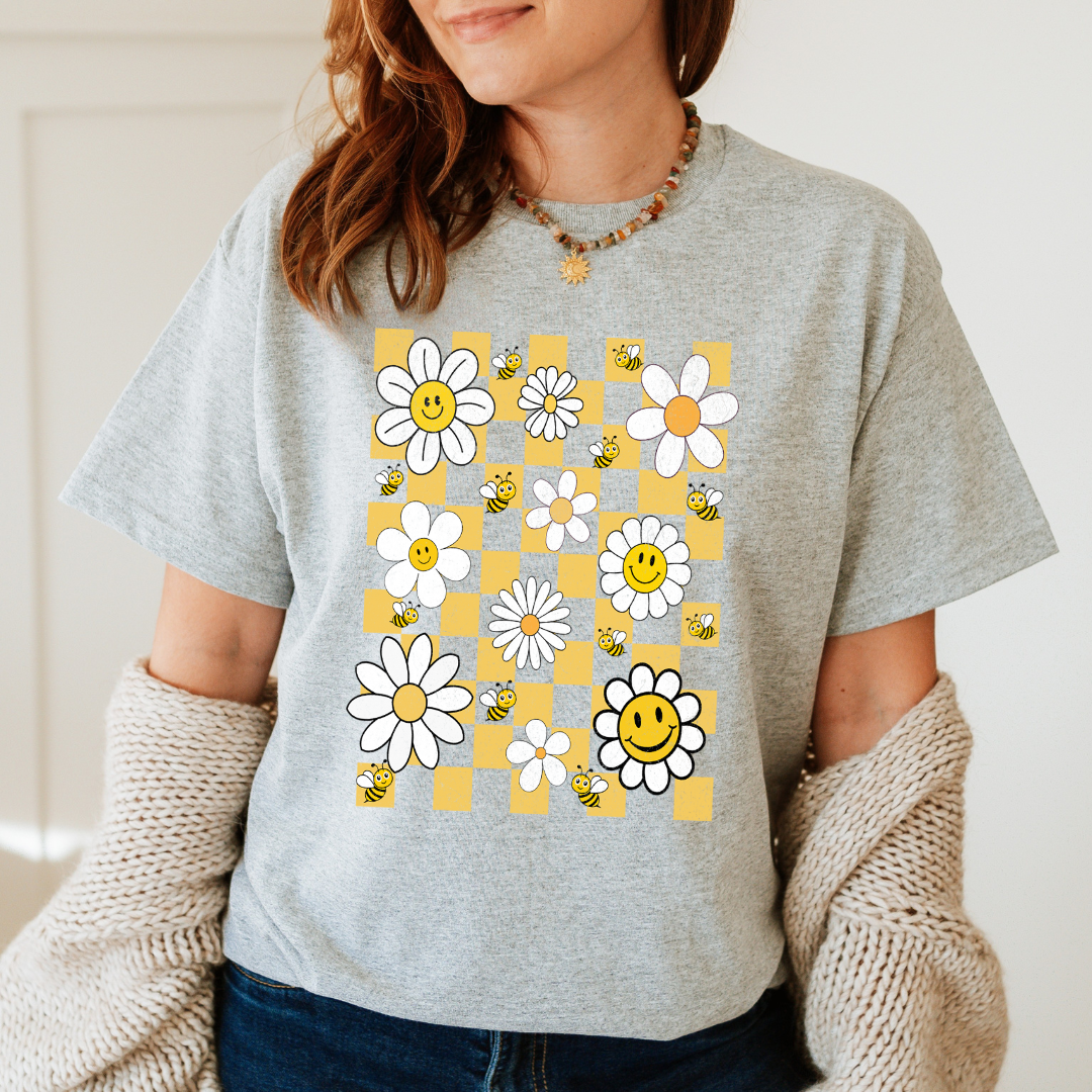 Femme portant un t-shirt beige style rétro avec une illustration de plusieurs marguerites blanches et des petites abeilles.