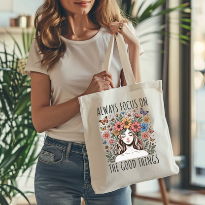 Sac fourre-tout en coton de couleur naturel de 15 x 16 pouces avec un design boho d'une femme entourée de fleurs colorées et de papillons, accompagnée du texte “Always Focus on the Good Things”.