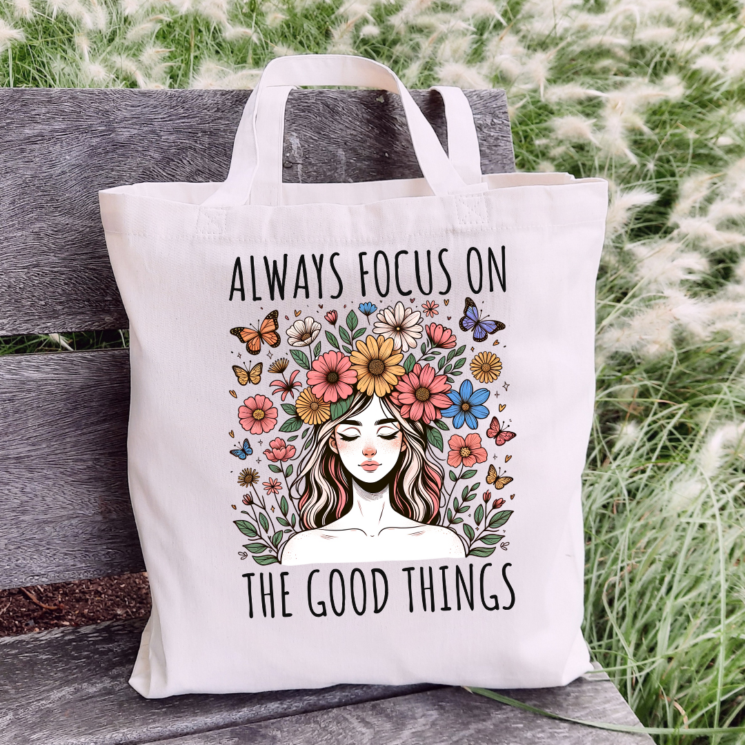 Sac fourre-tout en coton de couleur naturel de 15 x 16 pouces avec un design boho d'une femme entourée de fleurs colorées et de papillons, accompagnée du texte “Always Focus on the Good Things”.