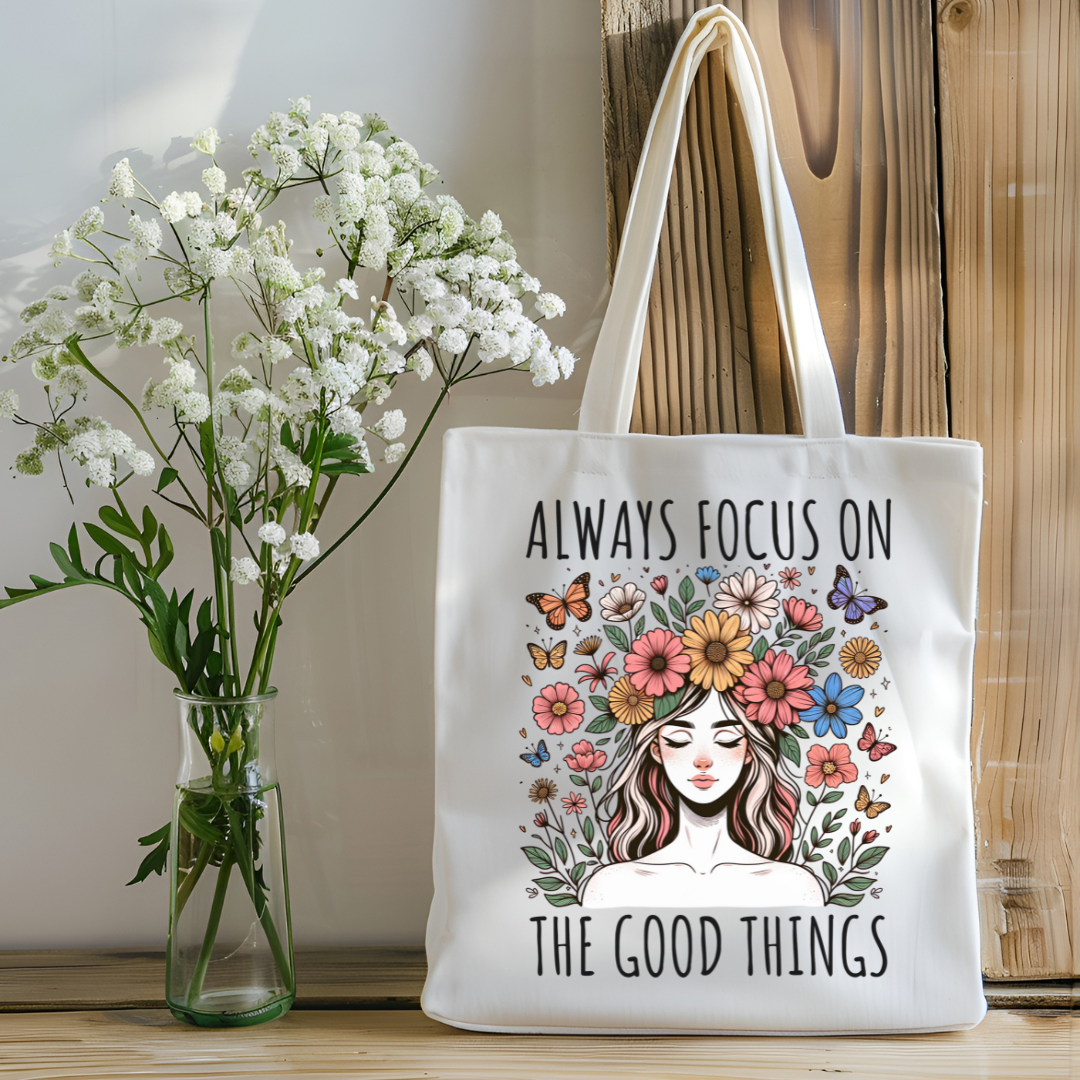 Sac fourre-tout en coton de couleur naturel de 15 x 16 pouces avec un design boho d'une femme entourée de fleurs colorées et de papillons, accompagnée du texte “Always Focus on the Good Things”.