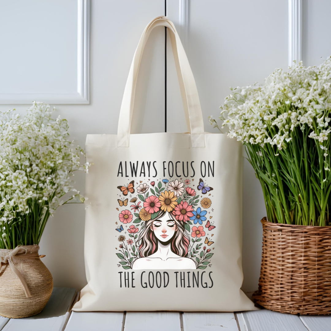 Sac fourre-tout en coton de couleur naturel de 15 x 16 pouces avec un design boho d'une femme entourée de fleurs colorées et de papillons, accompagnée du texte “Always Focus on the Good Things”.