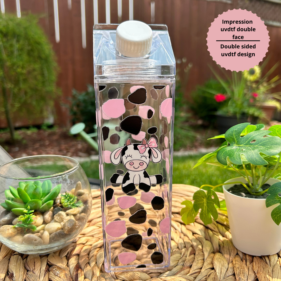 Bouteille transparente style carton de lait de 500 ml avec design de mignonne petite vache rose et noire, posée sur un plateau en osier à côté d'une plante.