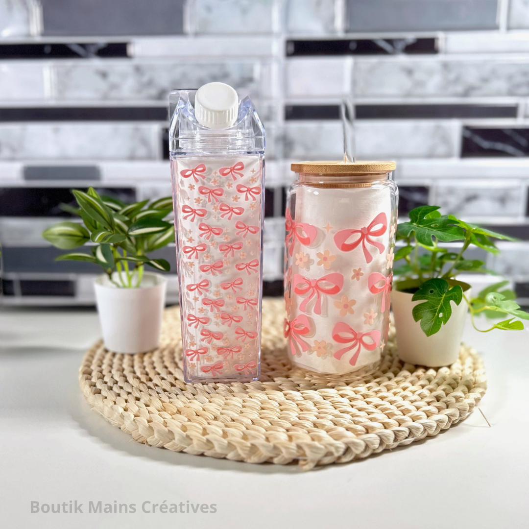 Duo de contenants à boisson style Coquette : une bouteille style carton de lait et un verre de 16 oz, tous deux décorés de motifs de boucles roses (pink bows) et de petites marguerites blanches sur un plateau de rotin.