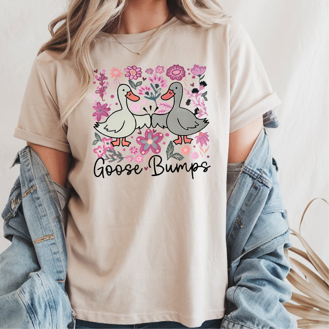 Femme portant un t-shirt beige avec une illustration d'oies se donnant le poing entourées de fleurs roses, texte "Goose Bumps", agencé avec une veste en jean.