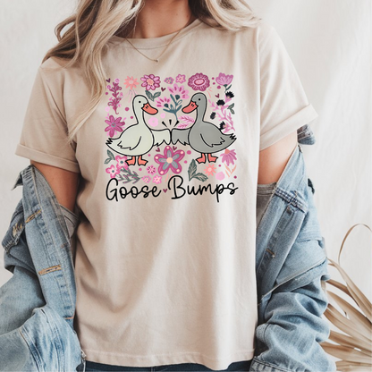 Femme portant un t-shirt beige avec une illustration d'oies se donnant le poing entourées de fleurs roses, texte "Goose Bumps", agencé avec une veste en jean.