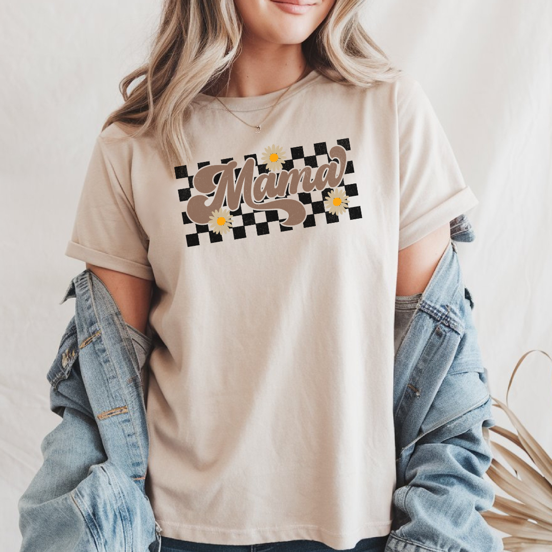 Femme portant un t-shirt beige avec le mot "Mama" en lettrage rétro à carreaux, décoré de marguerites blanches, agencé avec une veste en jean.