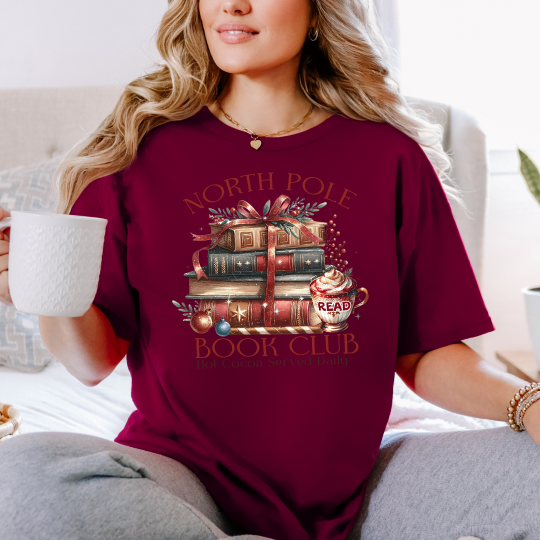 T-shirt Gildan Bourgogne avec un imprimé Livres de Noël empilés attachés d'un ruban rouge accompagnés d'une tasse de chocolat chaud. Ambiance cozy festive – parfait pour les fans de lecture !