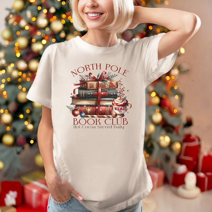 T-shirt Gildan Sable avec un imprimé Livres de Noël empilés attachés d'un ruban rouge accompagnés d'une tasse de chocolat chaud. Ambiance cozy festive – parfait pour les fans de lecture !