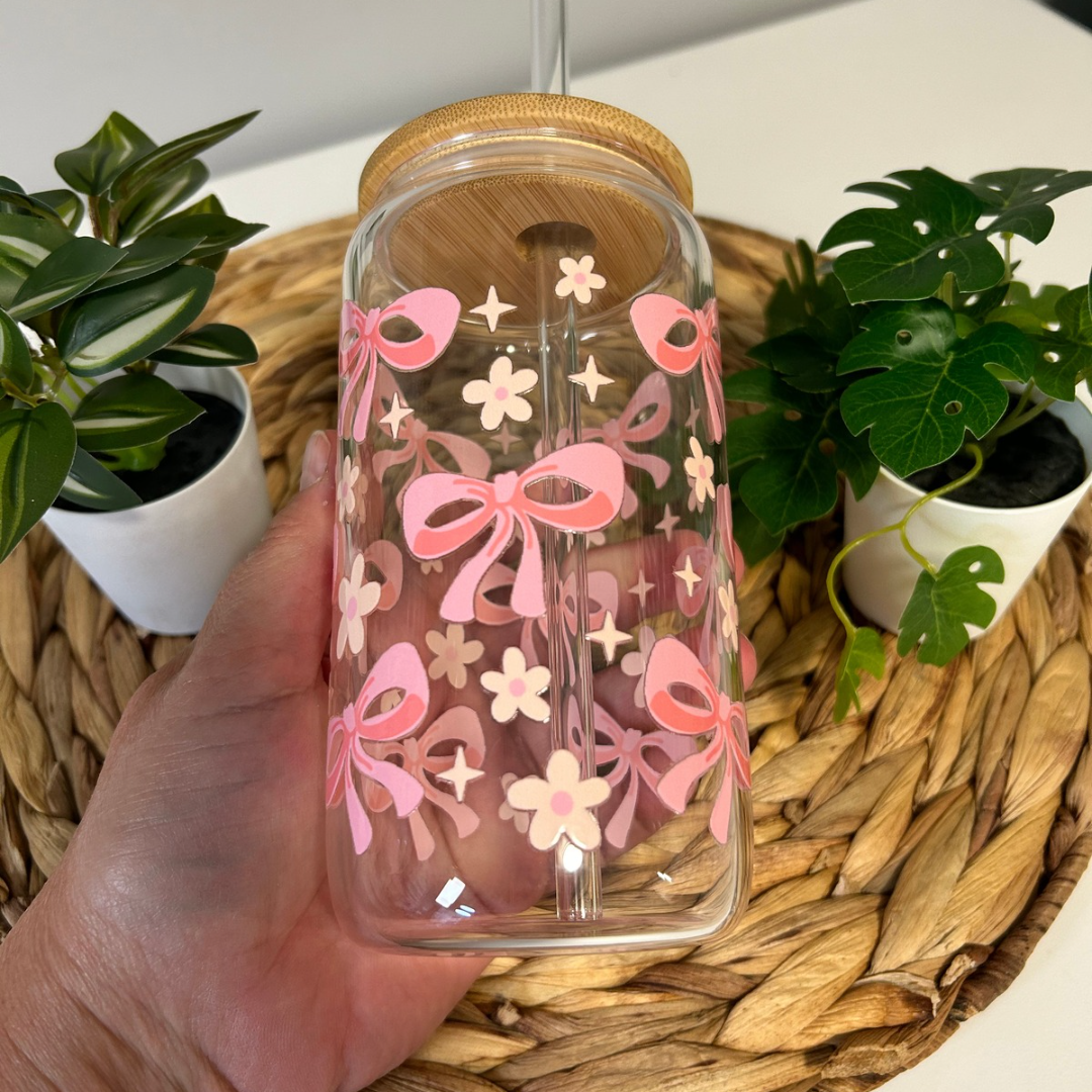 Verre à café glacé 16 oz avec motifs de petits nœuds roses et marguerites blanches, incluant un couvercle en bambou et une paille en verre, posé sur un support en bois.
