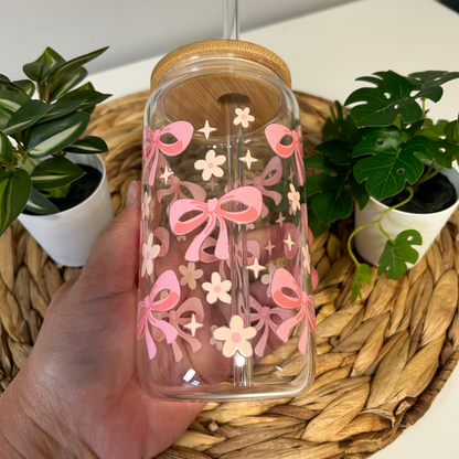 Verre à café glacé 16 oz avec motifs de petits nœuds roses et marguerites blanches, incluant un couvercle en bambou et une paille en verre, posé sur un support en bois.