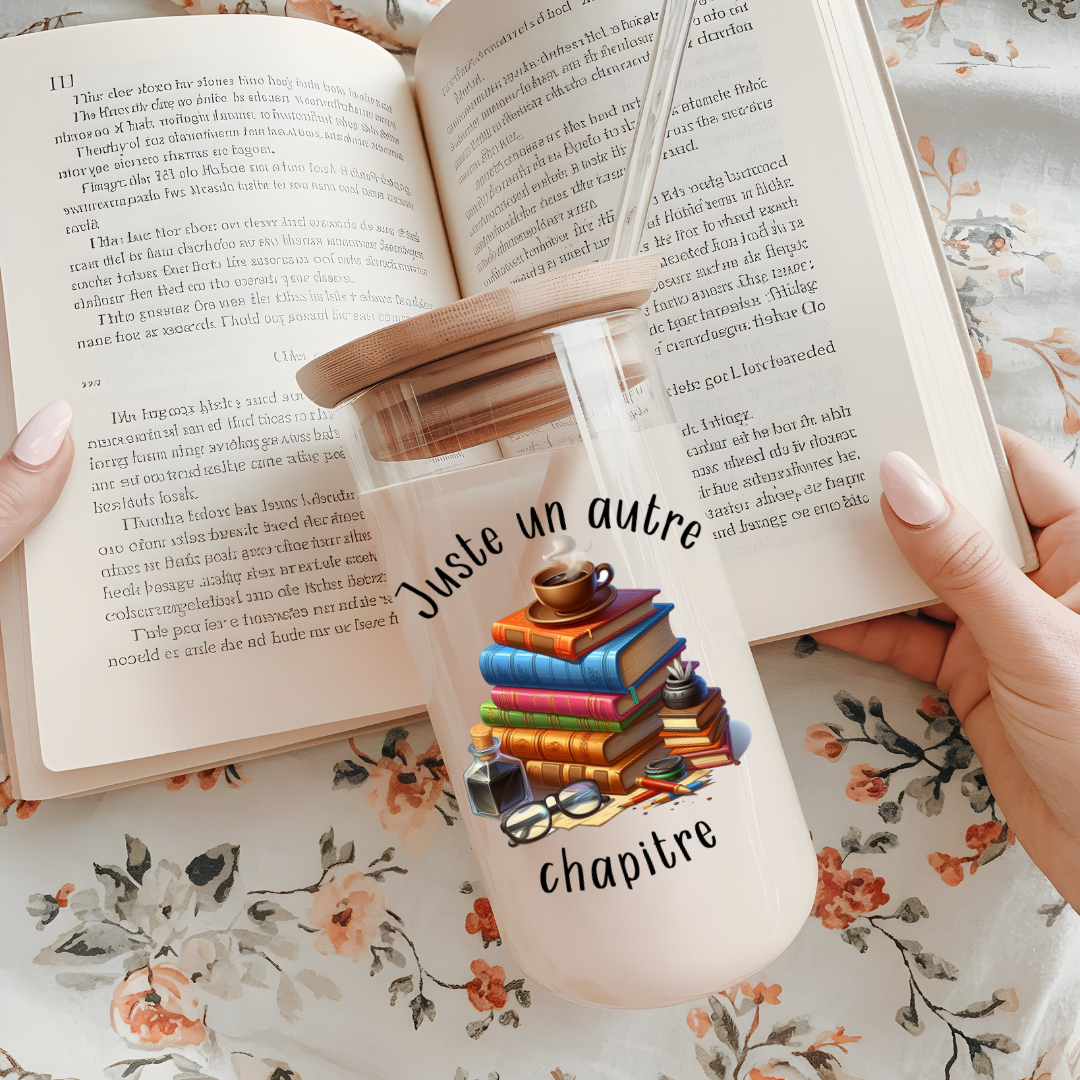 Verre à café glacé 16 oz avec motifs de livres empilés et tasses de café, incluant un couvercle en bambou et une paille en verre, posé sur un livre ouvert.